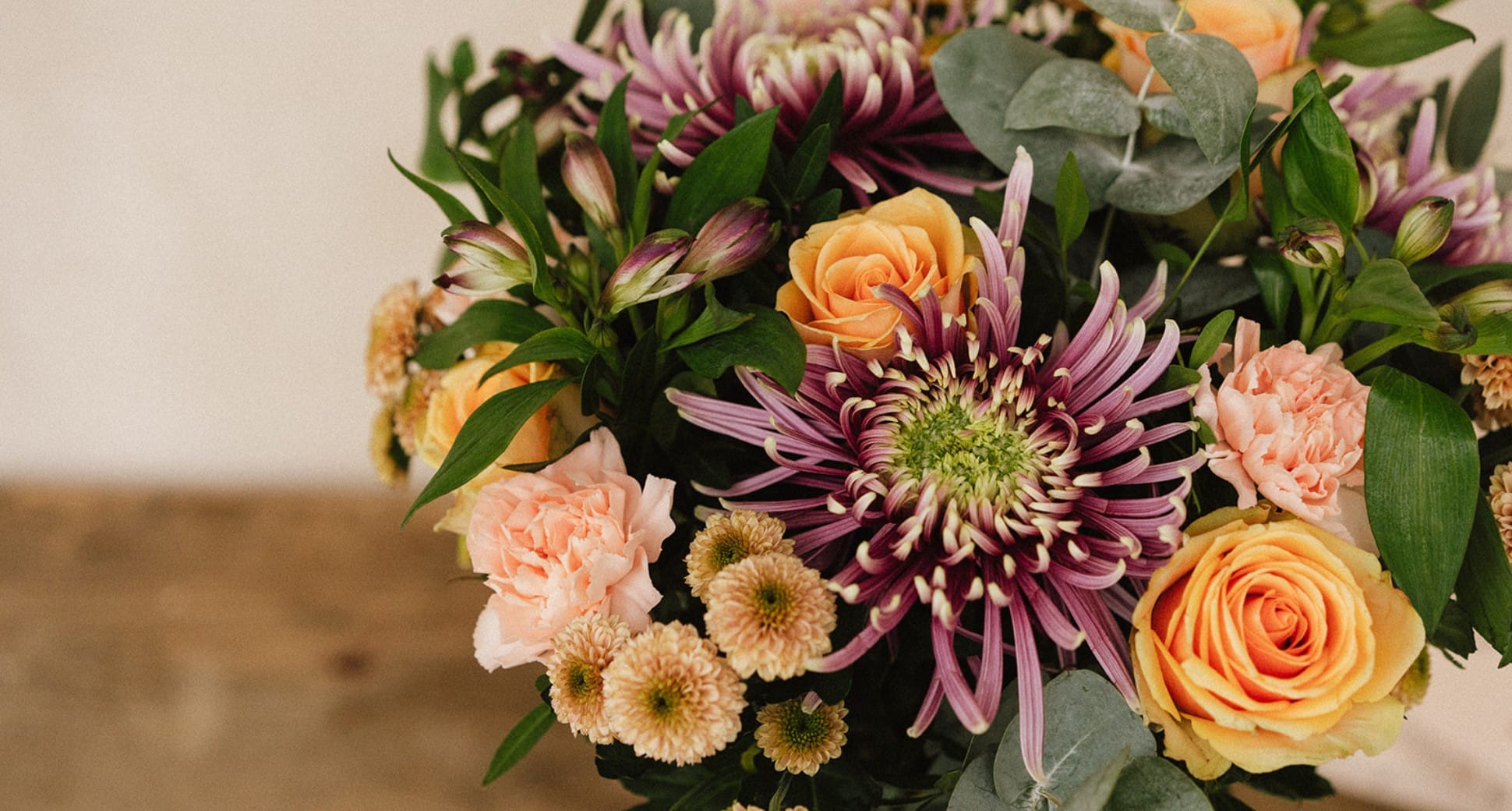 Bouquet de fleurs multicolores, principalement composé de roses jaunes, chrysanthèmes violets et petites fleurs blanches, disposé dans un vase, posé sur une table en bois. Arrière-plan flou beige.