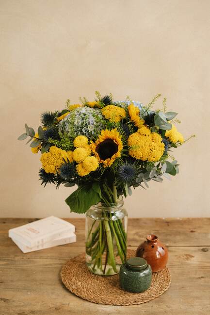 Le bouquet de fleurs jaune et vert repose dans un bocal en verre sur une table en bois, entouré d une napperon tressée, deux pots et un livre.