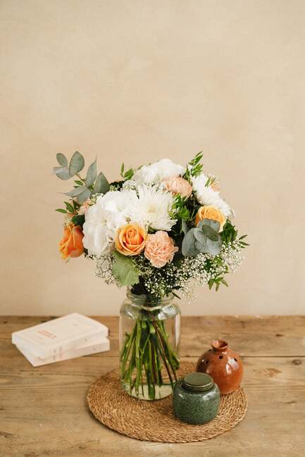 Un bouquet de fleurs, principalement blanches et oranges, repose dans un vase en verre sur une table en bois. À côté, deux pots en céramique et trois livres empilés.