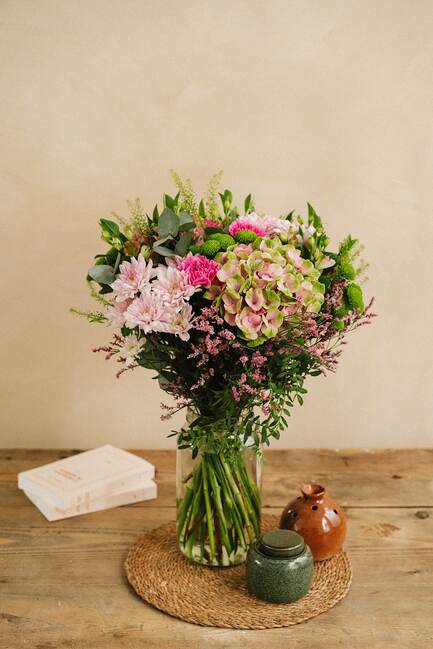 Bouquet de fleurs colorées dans un vase en verre, posé sur une table en bois avec des livres et deux petites jarres à côté.