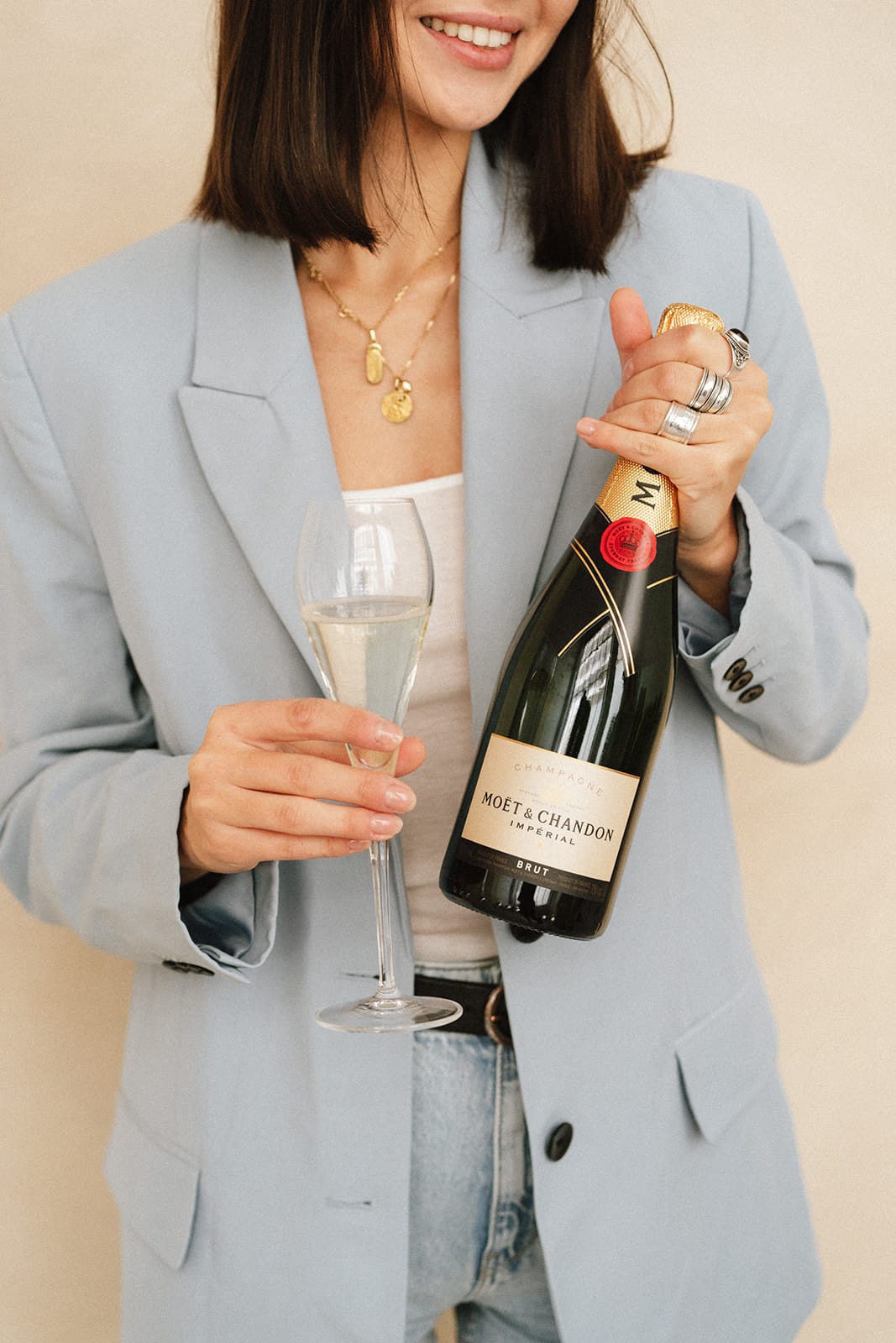 Une femme souriante, portant un blazer bleu, tient une bouteille de champagne Moët & Chandon ainsi qu un verre rempli du breuvage. Elle semble prête à célébrer.