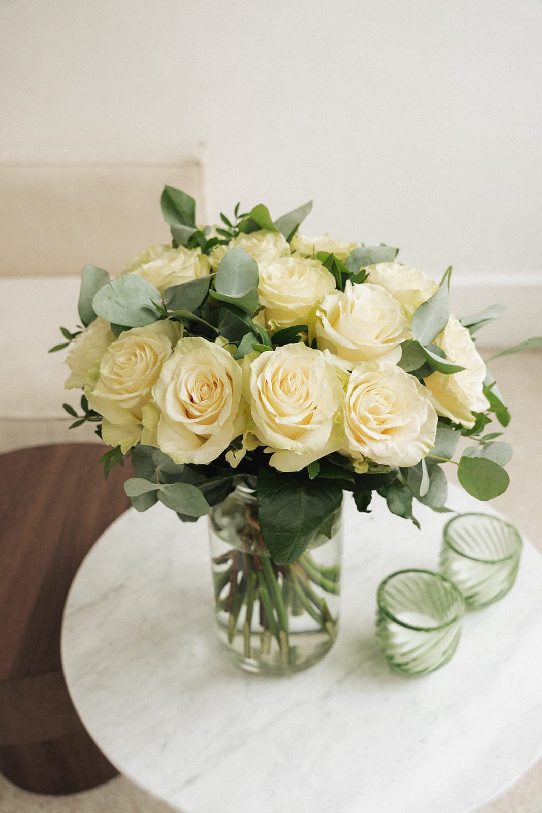 Un bouquet de roses blanches repose dans un vase en verre, placé sur une table en marbre, accompagné de deux bougies vertes en verre.