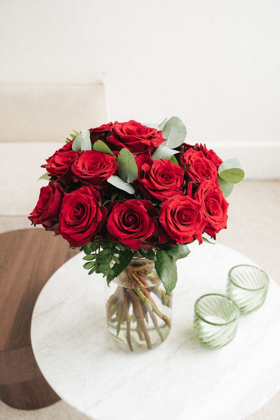 Un bouquet de roses rouges dans un vase en verre transparent, posé sur une table ronde en marbre blanc, à côté de deux verres verts.