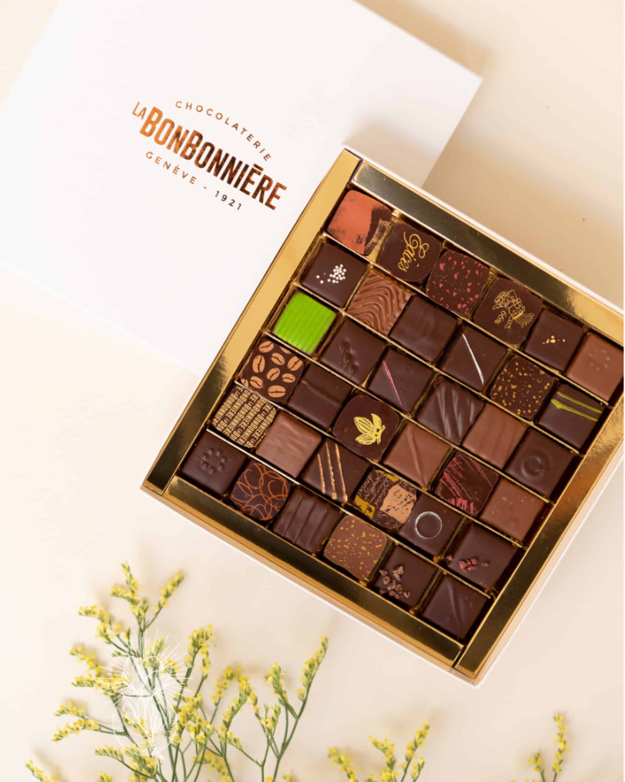 Une boîte ouverte de chocolats assortis, La Bonbonnière Chocolaterie Genève 1921, sur un fond beige avec des fleurs jaunes ornant le coin inférieur gauche.