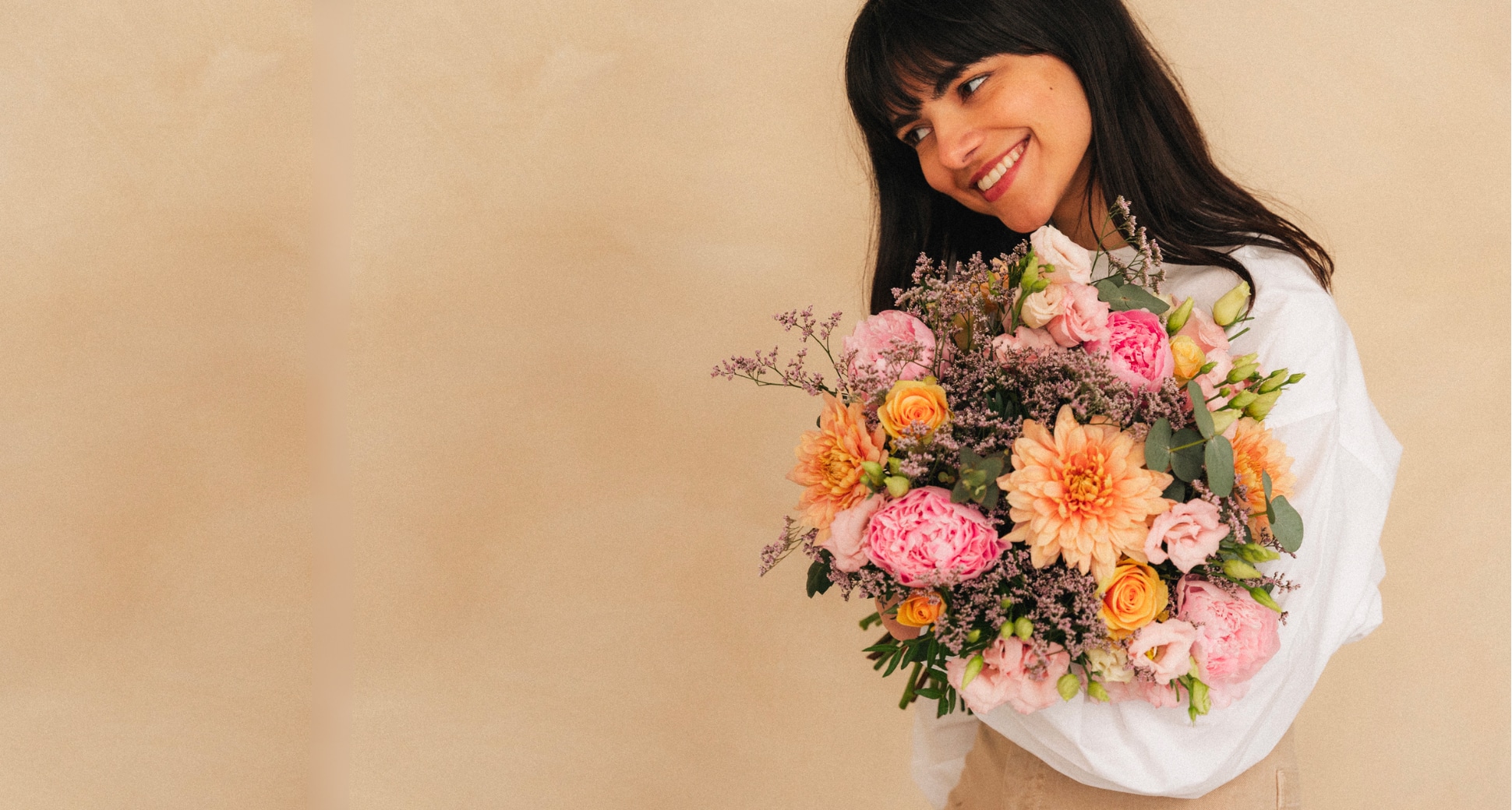 Femme souriante tenant un bouquet de fleurs multicolores, se tenant devant un fond beige uni.