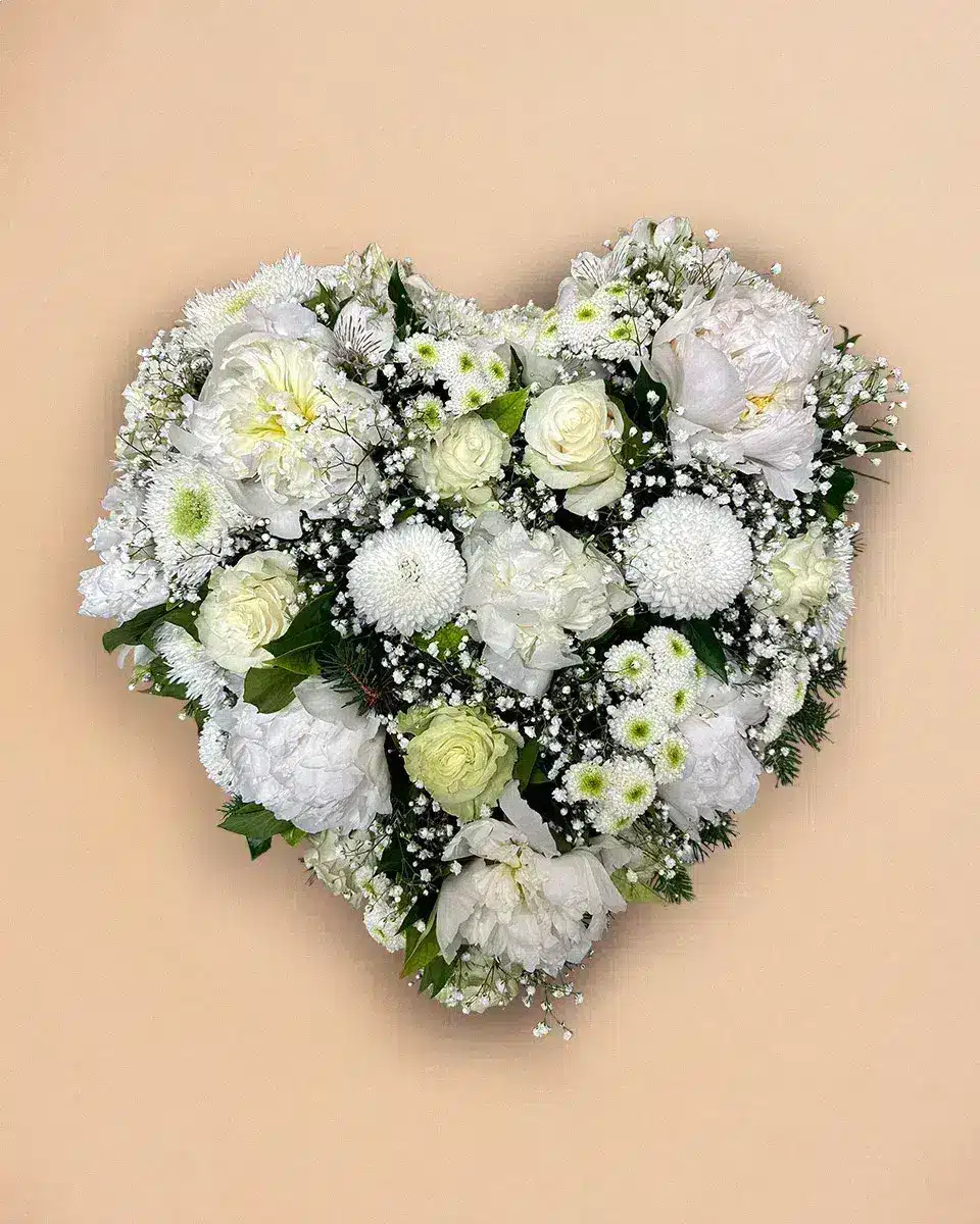 Livraison fleur de deuil : Un arrangement floral en forme de cœur comportant des roses blanches et d autres fleurs blanches, reposant sur un fond beige.