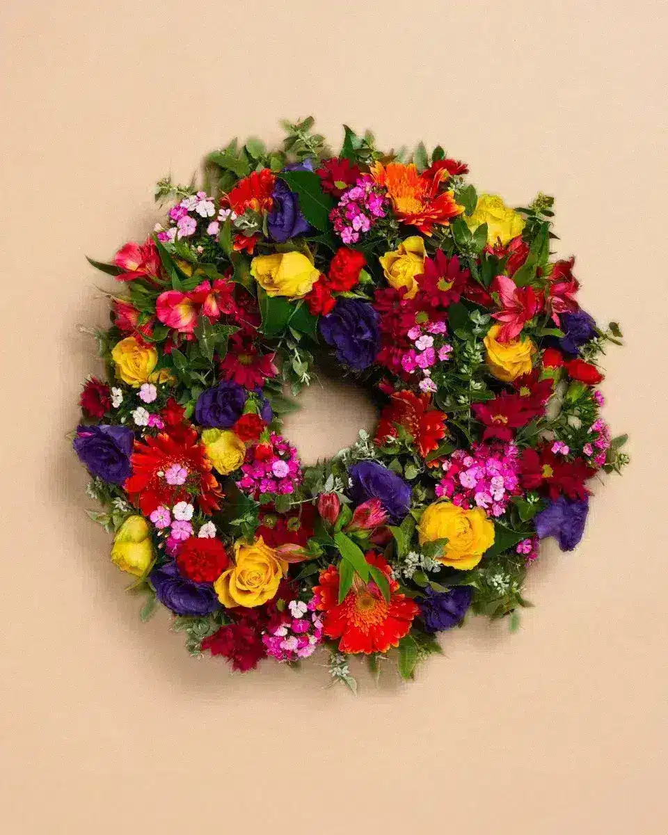 Livraison fleur deuil : Couronne de fleurs colorées disposées contre un mur beige, composées de roses jaunes, violettes, rouges et autres fleurs contextuelles.