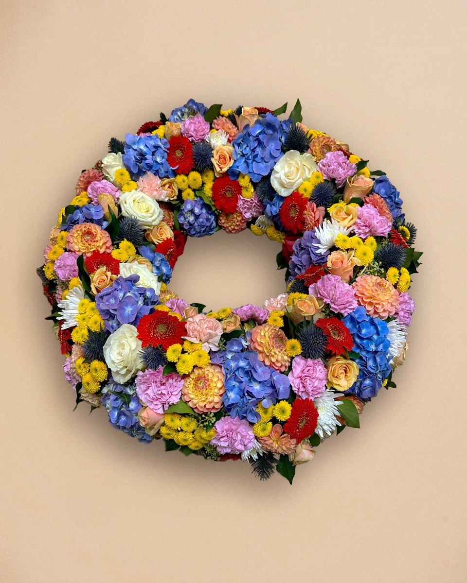 Livraison fleur deuil : Une couronne de fleurs multicolores (roses, bleues, jaunes, rouges) est accrochée à un mur beige.