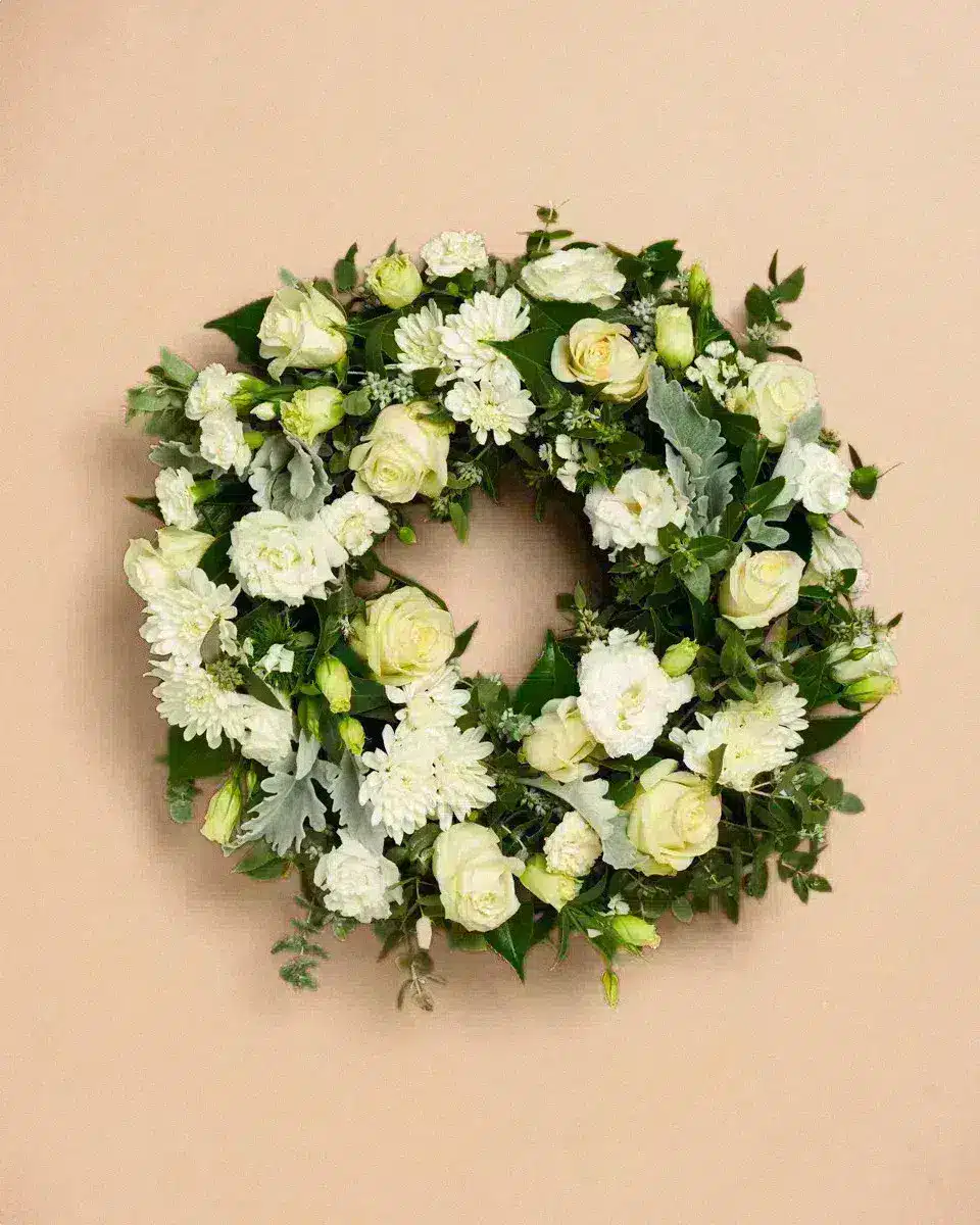 Livraison fleur deuil : Couronne de fleurs blanches et vertes accrochée sur un mur beige; les fleurs sont des roses, œillets et chrysanthèmes parmi des feuillages verts.