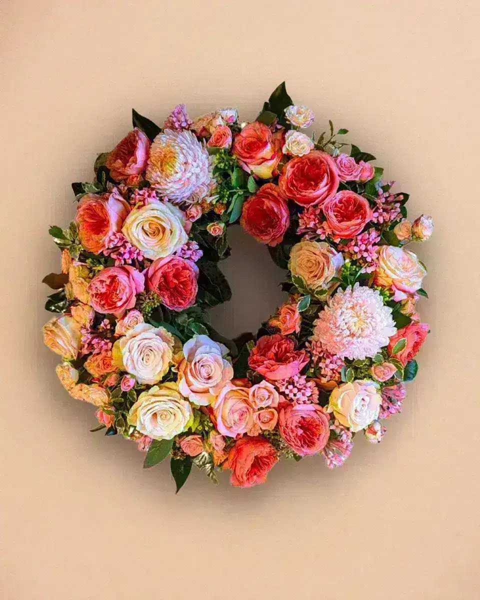 Livraison fleur deuil : Couronne florale ornée de roses rouges, roses et blanches, chrysanthèmes et feuillage vert, suspendue sur un mur beige.