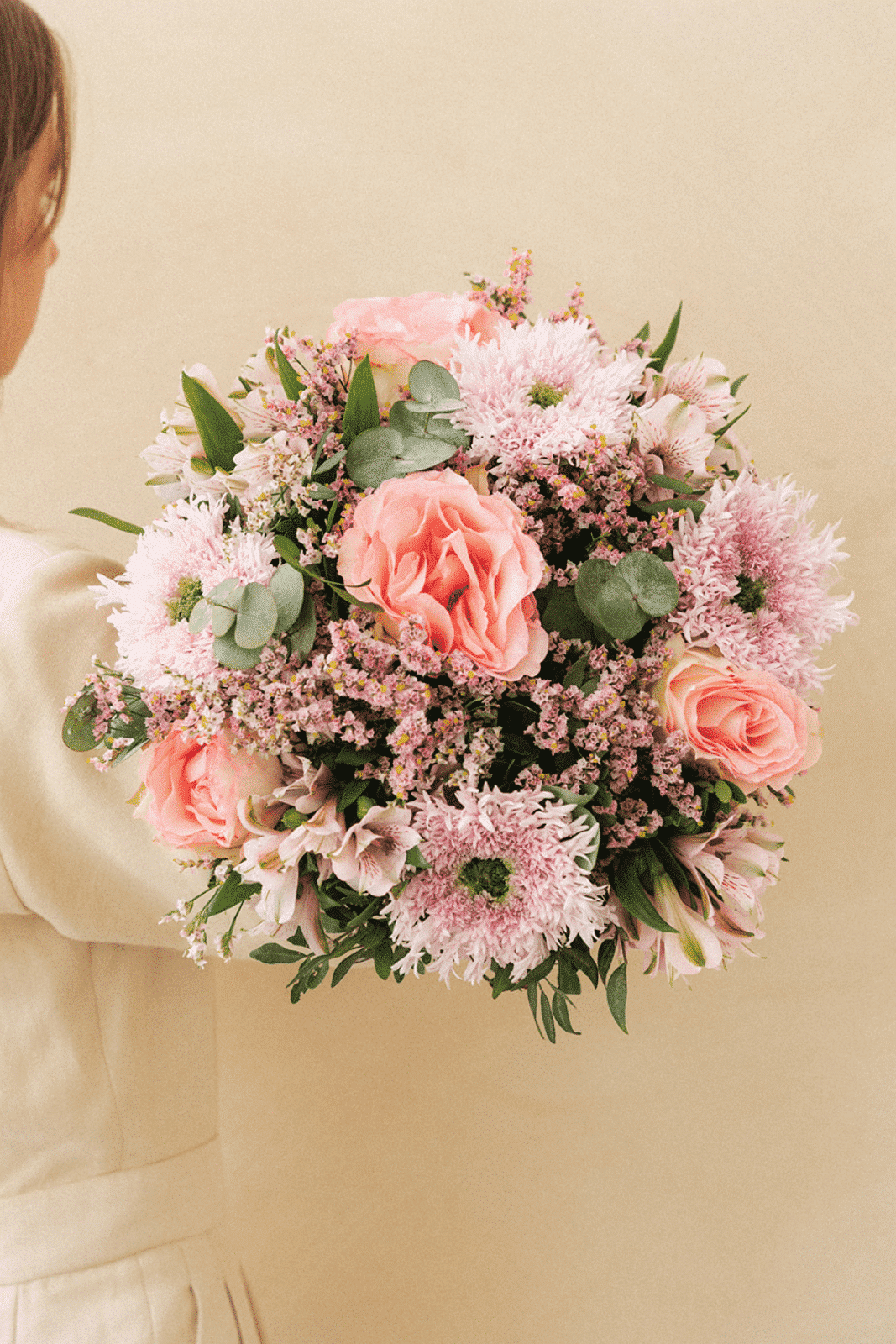 Bouquet de fleurs tenu par une personne, avec des roses roses, des chrysanthèmes mauves, des feuilles vertes, sur fond beige.