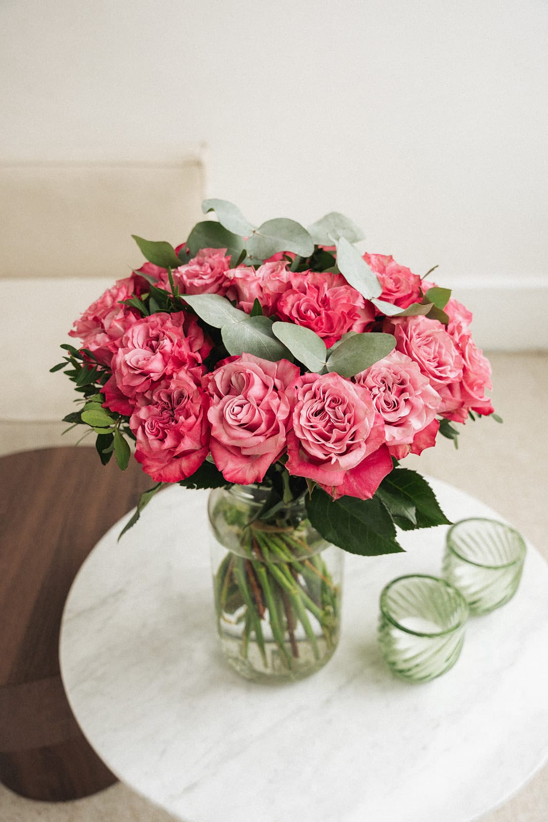 Un bouquet de roses roses est posé dans un vase en verre transparent sur une table en marbre, à côté de deux verres verts texturés, dans une pièce lumineuse et minimaliste.