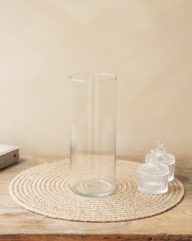 Vase en verre transparent reposant sur un dessous de plat tressé, entouré de deux pots en verre, sur une table en bois.