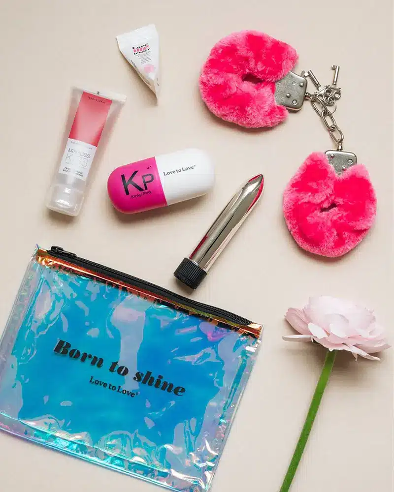 Divers objets sur une surface beige, dont menottes roses, tube de lubrifiant "MINUS 417 KISS", capsule rose et blanche avec "KP Love to Love", et une pochette holographique avec texte "Born to Shine".
