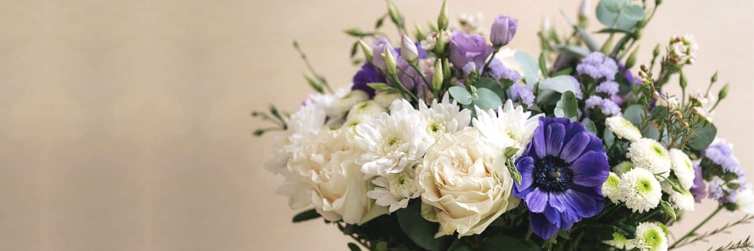 Bouquet de fleurs comportant des roses blanches, des chrysanthèmes blancs et des fleurs violettes, posé dans un vase, contre un fond beige uni.