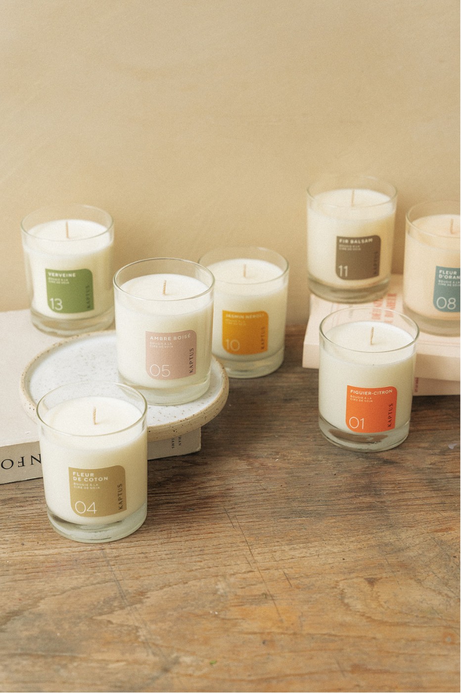 Sept bougies parfumées blanches dans des verrines transparentes, posées sur une table en bois, affichent les étiquettes
