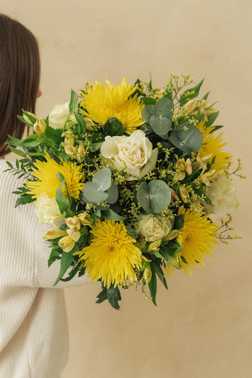 Un bouquet de fleurs jaunes et blanches est tenu par une personne en pull blanc, sur un fond neutre.