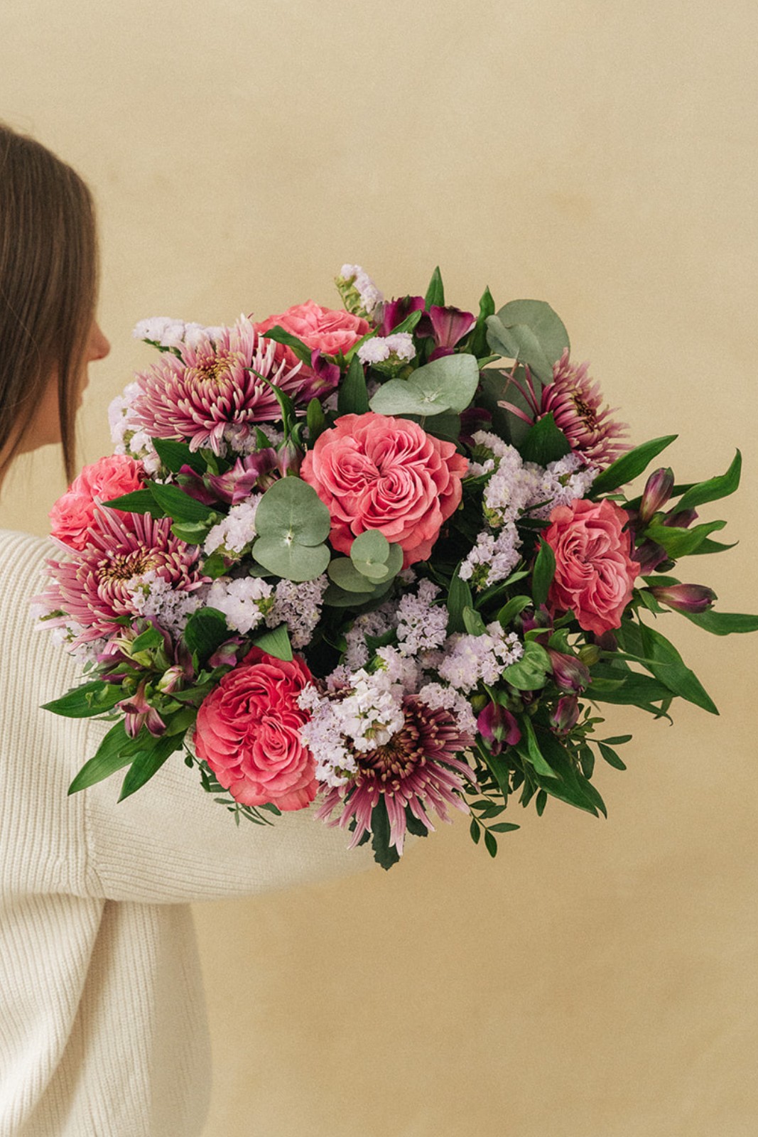 Un bouquet de fleurs colorées, principalement des roses et des chrysanthèmes, est tenu par une personne vêtue d un pull crème, dans un fond beige.